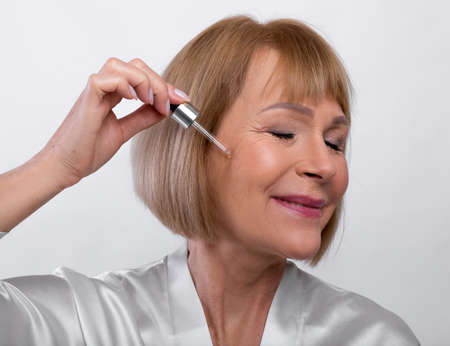 Portrait of happy senior woman in dressing gown applying face serum with dropper on light studio...