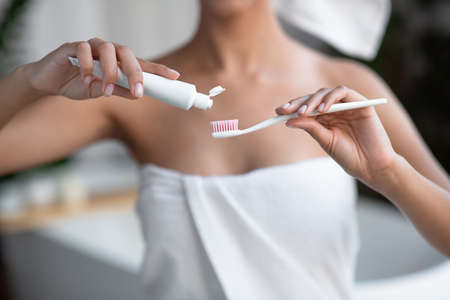 Morning personal oral hygiene, daily procedure and dental care. Millennial african american lady in towel squeezes toothpaste on toothbrush in bathroom interior, close up, copy space, croppedの写真素材