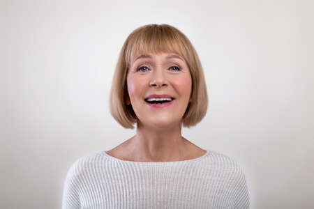 Portrait of happy elegant senior lady smiling, wearing sweater, looking at camera, posing over light...