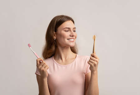 Smiling young woman holding eco bamboo and plastic toothbrush, choosing between different items for...