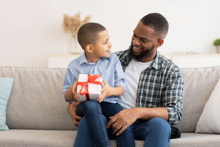 Black Daddy Congratulating Kid Son Giving Birthday Gift Box To Him Sitting On Sofa Together At Home. Family Holiday Celebration. B-Day Presents And Gifts Conceptの写真素材