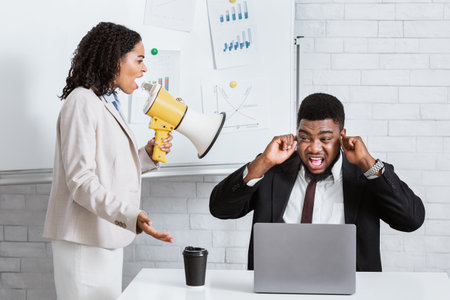 Enraged female boss with loudspeaker screaming at her scared subordinate at company office. Frightened male worker closing his ears, black businesswoman with megaphone yelling at him at workplaceの写真素材