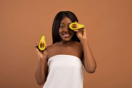 Happy beautiful black woman with long hair wrapped in towel covering eyes with half of avocado over beige studio background. Youth, beauty, anti-aging organic recipes, healthy diet conceptの写真素材
