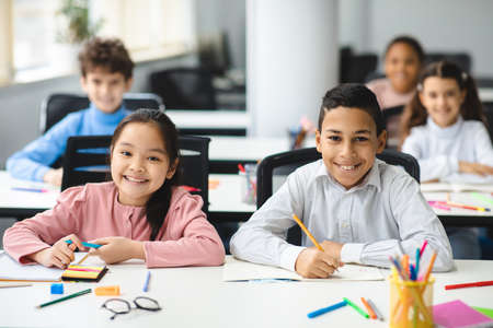 Education And Children Concept. Portrait of happy smiling diligent boy and girl sitting at table in classroom at elementary school with group of classmates in background and posing, looking at cameraの写真素材