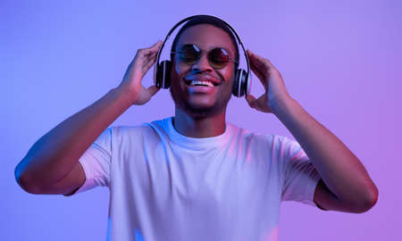Portrait Of Cheerful African American Guy In Wireless Headphones And Sunglasses In Neon Light, Happy Handsome Black Man In White T-Shirt Standing Over Purple Studio Background, Closeup With Free Spaceの写真素材