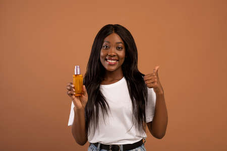 Pretty young black woman with long beautiful hair holding bottle with hair product, serum or oil, beige studio background, copy space. Hair care, healthy and beautiful hair conceptの写真素材