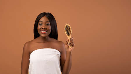 Attractive young black woman recommending wooden hair brush, posing wrapped in white towel with long shining fair hair holding brush over beige studio background, panorama with copy spaceの写真素材