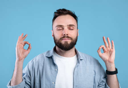 Calm young man meditating to relieve stress, finding inner peace and harmony on blue studio background. Bearded millennial guy practicing mindfulness and deep concentrationの写真素材