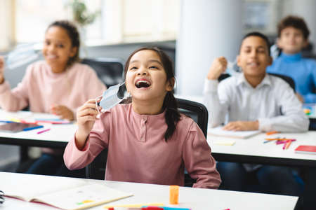 Multicultural group of children removing and taking off single-use surgical medical masks, sitting at table in classroom during school lesson and breathing fresh air. Studying And Health Care Conceptの写真素材