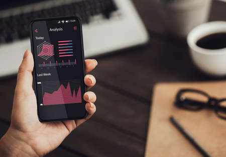 Woman using smartphone with online statistic and data analytics graphs and charts on screen while sitting at workplace in office, checking business information, creative collage, closeupの写真素材