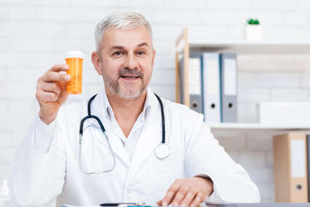 Drug treatment, prescription and doctors suggestion online. Friendly senior male in white coat with stethoscope holds jar of medicines and advises to clinic patient at video call in office, copy spaceの写真素材