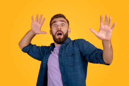 Scared Caucasian man making frightened gesture with his hands, trying to defend himself on orange studio background. Fearful young guy opening his mouth in shock, overwhelmed with emotionsの写真素材