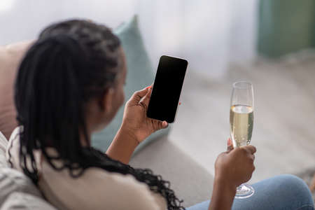 Unrecognizable black woman with glass of champagne using smartphone with blank screen, mockup, copy...