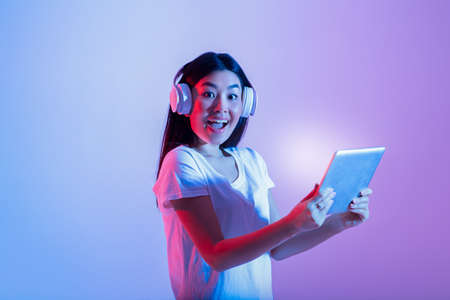 Human emotions of success, online win and modern gadgets. Surprised excited joyful young female with open mouth in headphones looks at camera and holds tablet with glowing screen, in neon, studio shotの写真素材