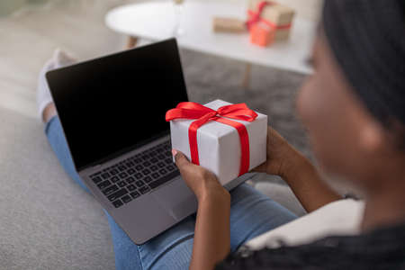 Unrecognizable black woman using laptop with empty screen, holding gift box, over shoulder shot, closeup. African american lady having birthday party online, celebrating with friends or familyの写真素材