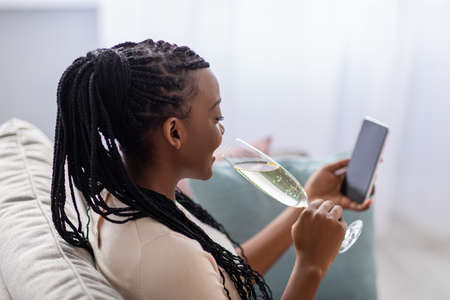 Pretty black woman with dreadlocks drinking champagne while using smartphone with blank screen, mockup, copy space. African american lady celebrating St. Valentines Day with her lover onlineの写真素材