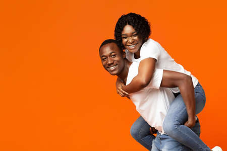 Funny black lovers young man and woman posing on orange studio background, copy space. Cheerful african american woman piggybacking on her man, standing by empty space for advertisementの写真素材