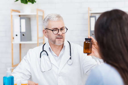 Medications for patient treatment and recommendations from doctor. Serious adult male in white coat and glasses shows jar of medicines to lady patient at table with sanitizer in office interiorの写真素材