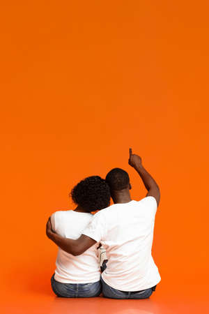 Rear view of black couple cuddling while sitting on floor together over orange studio background. African american man hugging his girlfriend and pointing at copy space, watching starsの写真素材