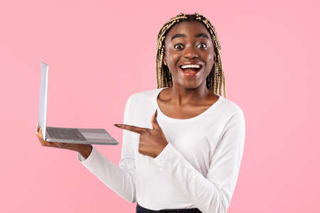 Portrait of excited smiling african american young woman holding laptop and pointing at gadget screen, looking at camera, standing isolated over pink studio background. People, Technologyの写真素材