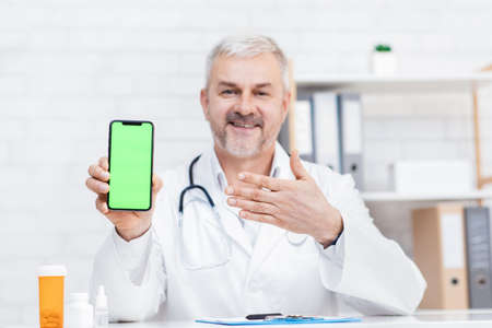 Modern medical app for online chat with doctor. Cheerful adult man in coat with stethoscope sits at...
