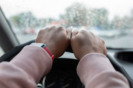Difficult road in bad rainy weather. Hands of young african american or mixed race lady with smartwatch on steering wheel, lady drives car in city, view of window with drops, pov, copy space, close upの写真素材