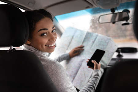 Lady enjoying comfortable auto journey sitting on driver seat. Happy cute millennial african american lady holding map and smartphone with blank screen, turns and looks at passenger from behindの写真素材