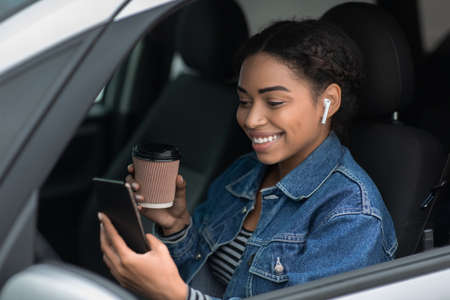 Portrait of young and cheerful woman with coffee cup sits in car in city on break. Smiling pretty...