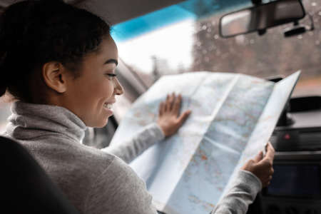 Young woman travel alone with map in car in new town. Smiling beautiful millennial african american...