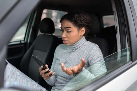 Travel problems and get lost in new city and looking for road, sitting in car. Upset confused excited young african american woman with map looks in smartphone and cannot find way, empty spaceの写真素材