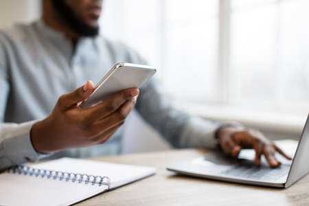 Business Application. Black Businessman Using Phone Working On Laptop At Workplace Indoors. Mobile...