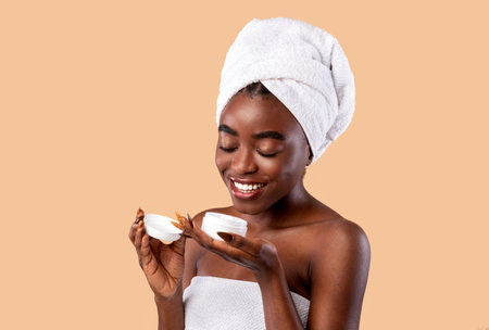 Skin Care Concept. Portrait of beautiful young african american woman holding open cream bottle looking inside. Smiling black lady wearing white towel on head isolated over studio backgroundの写真素材
