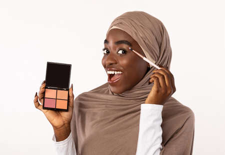 Everyday Nude Makeup. Portrait of beautiful excited african american muslim woman wearing hijab applying cosmetics, holding brush and eyeshadow palette and showing it to camera, studio backgroundの写真素材