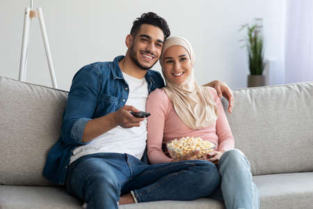 Happy muslim family of two watching TV together at home, hugging and eating popcorn, copy space....