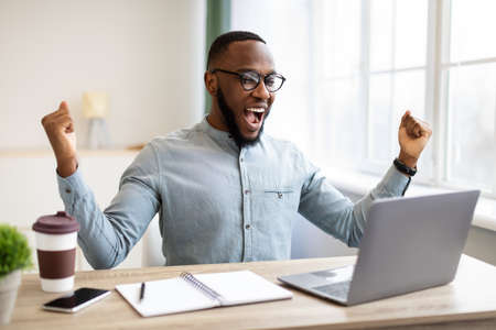 Business Success. African American Businessman At Laptop Shaking Fists And Shouting In Joy Celebrating Career Promotion Sitting At Workplace In Modern Office. Successful Entrepreneurship Conceptの写真素材