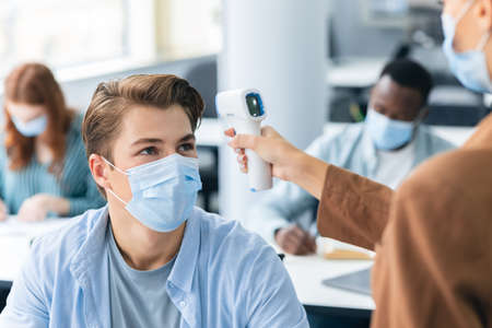 Woman using digital medical contactless infrared electronic thermometer, measuring and checking body temperature, screening male student in protective mask, preventing contagious diseases spreadの写真素材