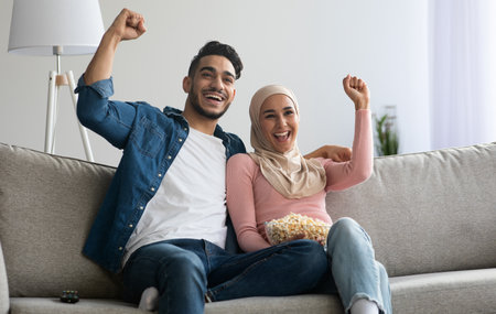 Emotional muslim couple football fans watching game on TV, raising hands up, celebrating success of their team, happy arab man and woman in hijab sitting on couch at home, eating popcornの写真素材
