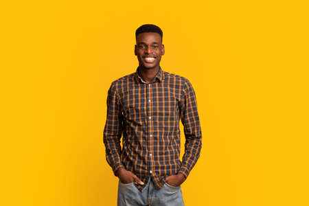 Portrait Of Young Handsome Black Guy In Checkered Shirt Over Yellow Background, Cheerful Millennial African American Man Standing With Hands In Pockets, Looking At Camera And Smiling, Copy Spaceの写真素材