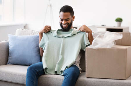 Delivery Concept. Happy Black Man Unpacking Cardboard Box Holding New T-Shirt Sitting On Sofa At Home. Customer Receiving His Order. E-Commerce And Online Shopping, Purchase And Clothes Shipmentの写真素材