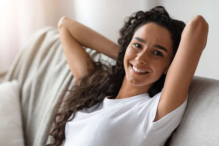 Closeup of carefree young woman with dark curly hair resting at home alone in cozy atmosphere on weekend, leaning back on sofa and relaxing, smiling at camera, copy space, living room interiorの写真素材