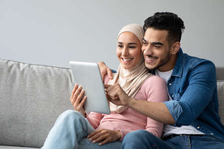 Excited arab guy showing something his girlfriend on pad. Happy middle-eastern muslim couple enjoying time together at home, using digital tablet, websurfing or shopping online, copy spaceの写真素材