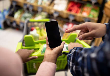 Unrecognizable black couple holding smartphone with empty screen at supermarket, mockup for your mobile app or website. African American guy and lady with cellphone shopping at mallの写真素材