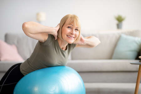 Smiling senior woman doing exercises with fitness ball at home, space for text. Mature Caucasian lady working out her back muscles, training with sports equipment during coronavirus lockdownの写真素材