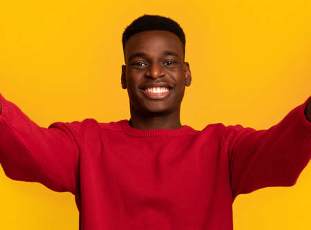 Selfie Time. Cheerful young african american guy making self-portrait and smiling at camera, positive black man taking photo standing isolated over yellow background, closeup shot with free spaceの写真素材