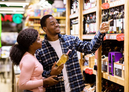 Happy black man and his girlfriend buying wine at liquor department of supermarket. Lovely African American couple shopping for alcoholic drinks at huge mall togetherの写真素材