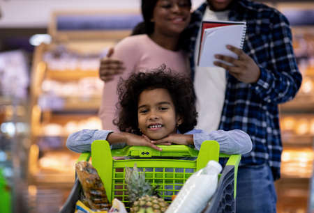 Adorable black girl with full shopping cart and her parents checking grocery list at modern supermarket. Millennial African American family with cute child doing weekly purchases at mallの写真素材