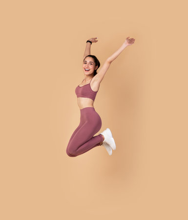 Full Length Portrait Of Happy Excited Smiling Young Woman In Sports Bra And Leggins Jumping Up And Raising Arms Up Isolated On Pastel Background, Celebrating Success, Studio Shot, Copy Space.の写真素材