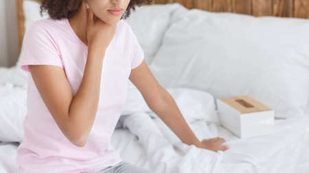 Sore throat, pain, disease and viral infections. Desperate african american millennial woman sits on white bed and presses hand to throat in cozy interior of bedroom, panorama, free space, croppedの写真素材