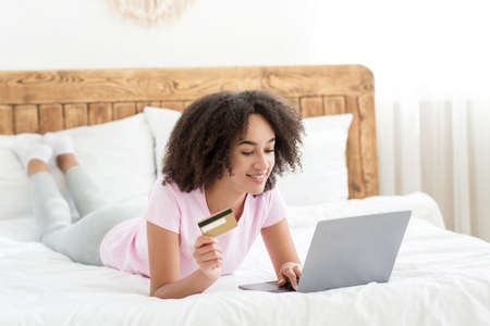 Online shopping on bed and good offer at home. Smiling millennial cute african american female lies on bed, looks at laptop, holds credit card, in cozy bedroom interior in daylight, free spaceの写真素材