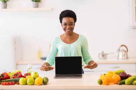 African American Lady Showing Blank Laptop Screen For Cooking Website Advertisement Posing In Modern Kitchen At Home, Smiling To Camera. Weight Loss And Healthy Nutrition Blog. Mockupの写真素材
